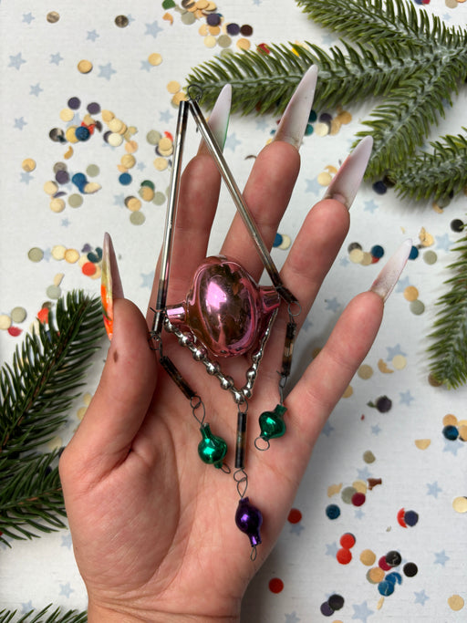 A hand holding a small, colorful ornament with a metallic frame, surrounded by festive decorations like confetti and pine branches.