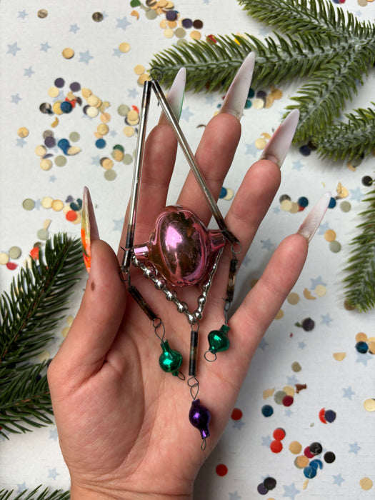 A hand holding a small, colorful ornament with a metallic frame, surrounded by festive decorations like confetti and pine branches.