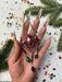 A hand holding a small, colorful ornament with a metallic frame, surrounded by festive decorations like confetti and pine branches.