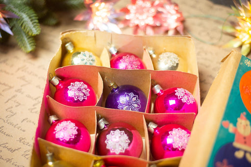 A cardboard box filled with colorful Christmas ornaments, including pink and purple ornaments with intricate designs.
