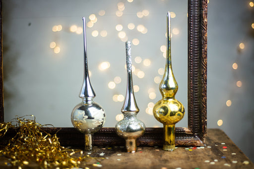 three decorative ornaments, including a silver cone, a gold cone, and a gold ball, arranged on a wooden surface. The ornaments are placed in front of a dark wooden frame, which has a blurred background with twinkling lights.