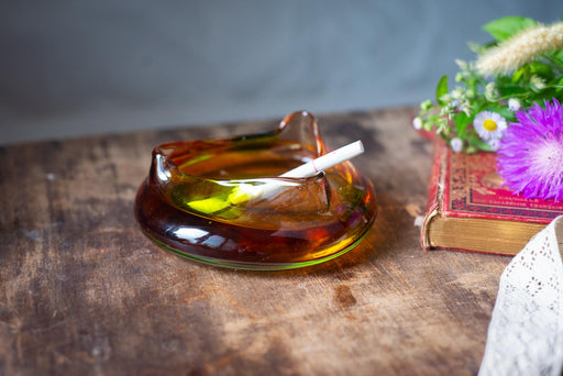 A glass ashtray containing a lit cigarette rests on a wooden surface, with a book and flowers nearby.
