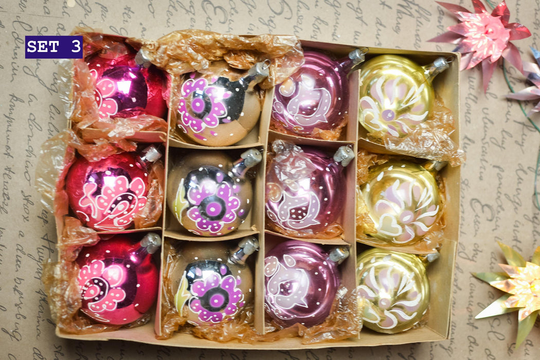 A set of colorful glass ornaments in a cardboard box, with various designs and colors including pink, purple, yellow, and green.