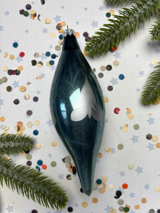 A blue glass ornament with a white feather design is surrounded by pine branches, confetti, and small star-shaped decorations on a white background.