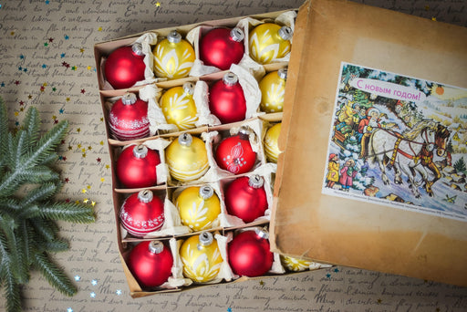 A cardboard box filled with red and yellow Christmas ornaments, with a handwritten note attached to the side of the box.