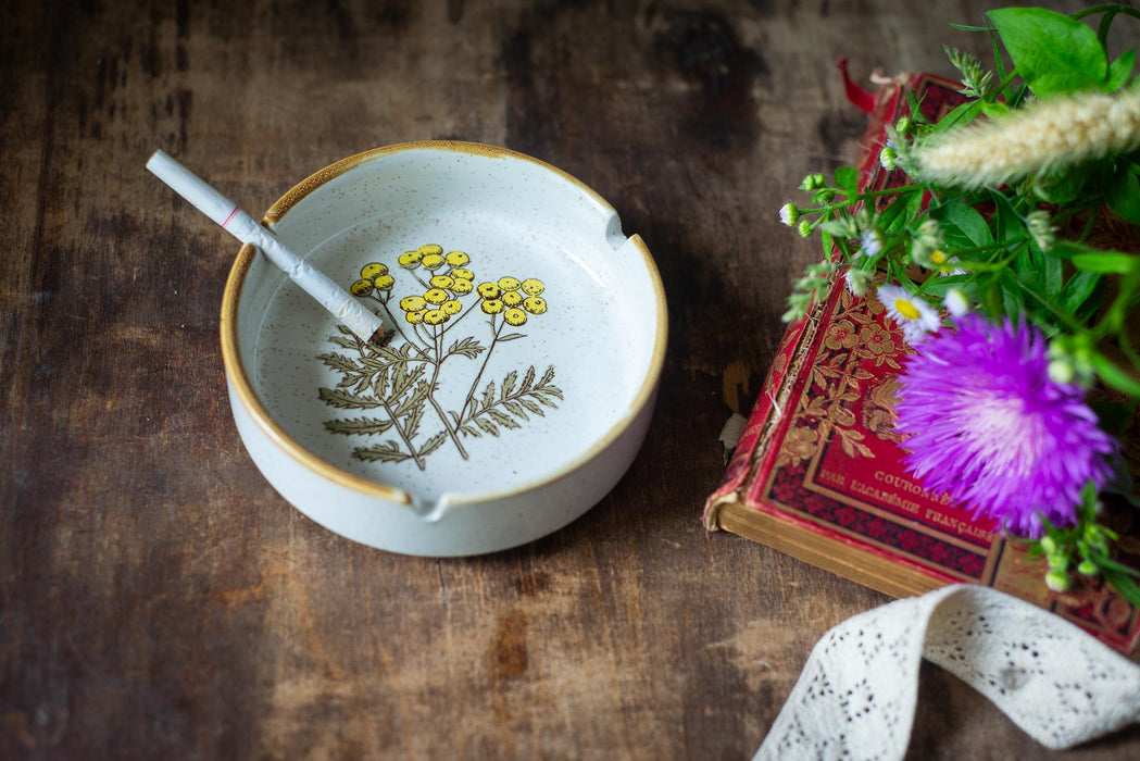 a white ceramic ashtray with a floral design, a lit cigarette, a purple flower, and a red book with gold lettering on a wooden surface.