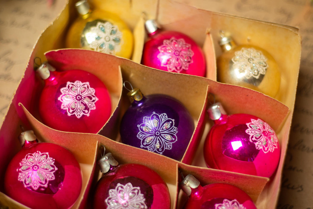 A cardboard box containing nine Christmas ornaments, with a mix of red, yellow, and purple colors.