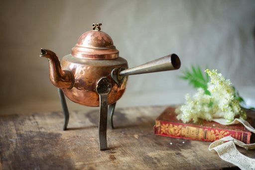 An antique copper teapot with a spout and handle sits on a wooden surface, accompanied by a bouquet of white flowers and an old book.