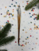 a metallic ornament with a long, curved shape, surrounded by colorful confetti and pine branches.