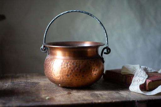 A copper-colored pot with a black handle sits on a wooden surface, accompanied by a white lace doily and a book.
