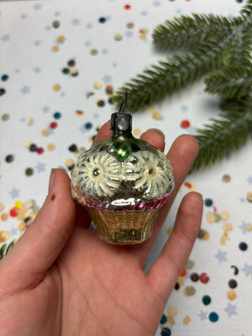 A hand holding a small, ornate glass ornament with a floral design, against a background of confetti and a pine tree branch.