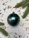 A shiny, black ornament with a cracked surface is surrounded by colorful confetti and a sprig of greenery, creating a festive and whimsical holiday scene.