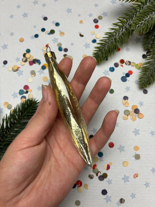 A hand holding a gold ornament against a background of colorful confetti and a pine tree branch.