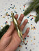 A hand holding a gold ornament against a background of colorful confetti and a pine tree branch.