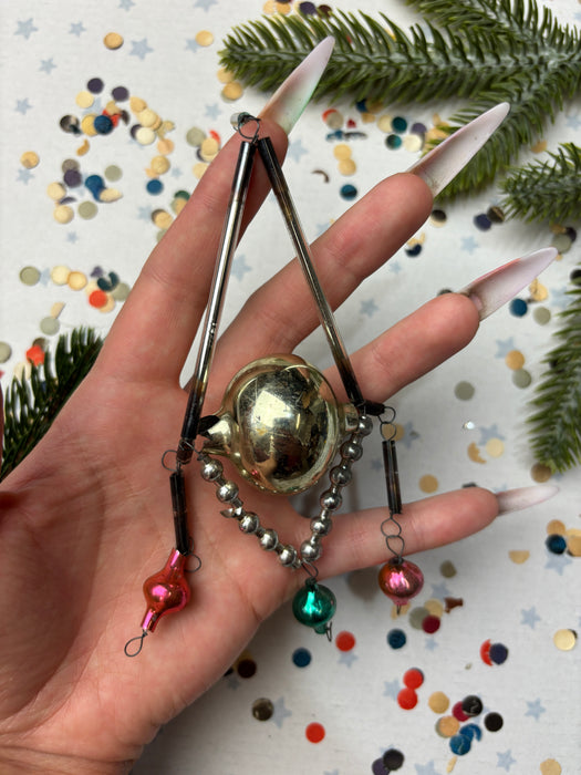 A hand holding a collection of ornaments, including a small gold ball, a silver ball, and a pink ball, against a background of scattered confetti and pine branches.