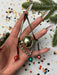 A hand holding a collection of ornaments, including a small gold ball, a silver ball, and a pink ball, against a background of scattered confetti and pine branches.