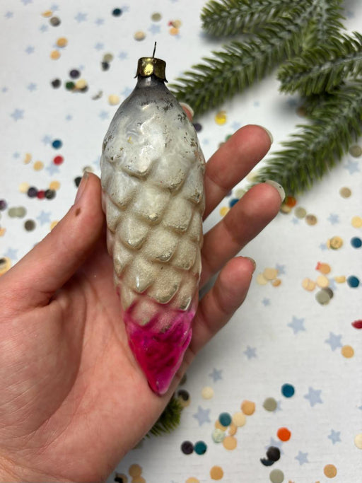 A hand holding a decorative ornament with a pink tip, against a background of colorful confetti and a pine tree branch.