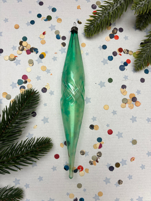 a green glass ornament with a twisted shape, surrounded by colorful confetti and pine branches.