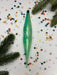 a green glass ornament with a twisted shape, surrounded by colorful confetti and pine branches.