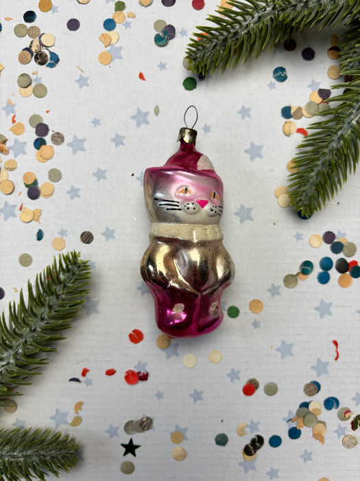 A pink and gold ornament with cat faces is surrounded by confetti and pine branches on a white background.