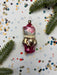 A pink and gold ornament with cat faces is surrounded by confetti and pine branches on a white background.
