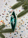 a green glass ornament with a white design, surrounded by colorful confetti and pine branches.
