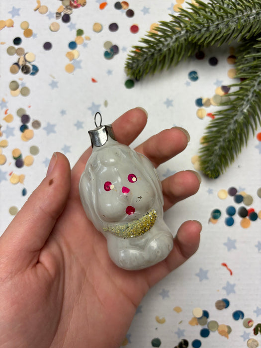 A hand holding a small white ornament with red and gold accents, surrounded by colorful confetti on a white background.