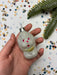 A hand holding a small white ornament with red and gold accents, surrounded by colorful confetti on a white background.