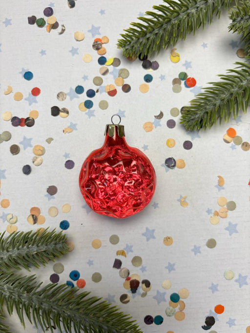 A red Christmas ornament is surrounded by colorful confetti and green pine branches on a white background.
