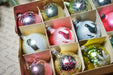 A cardboard box filled with a variety of colorful Christmas ornaments, including glass baubles, baubles with animal designs, and other festive decorations.