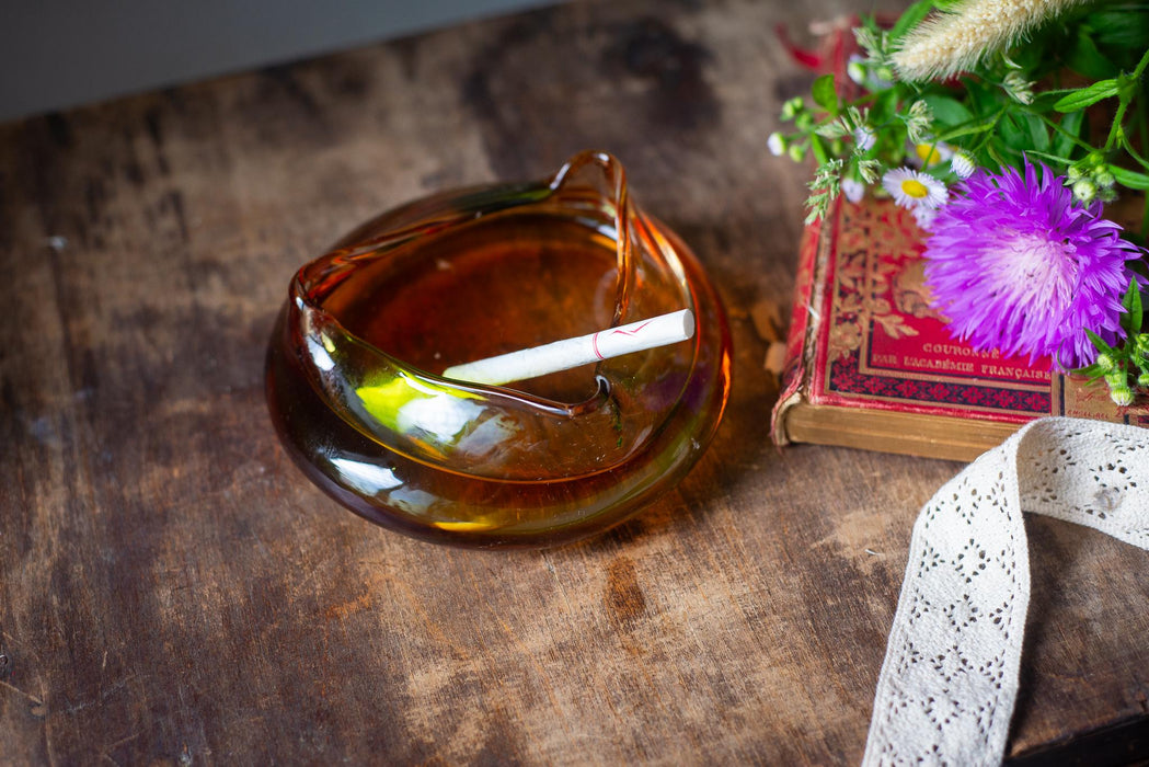 An amber glass ashtray with a lit cigarette rests on a wooden surface, accompanied by a book and a bouquet of purple flowers.