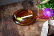 An amber glass ashtray with a lit cigarette rests on a wooden surface, accompanied by a book and a bouquet of purple flowers.