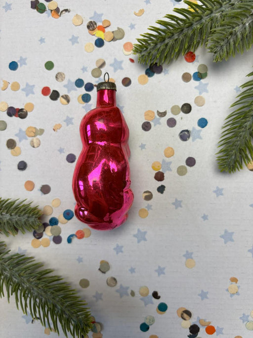 A shiny red ornament is placed on a white background, surrounded by colorful confetti and green pine branches.