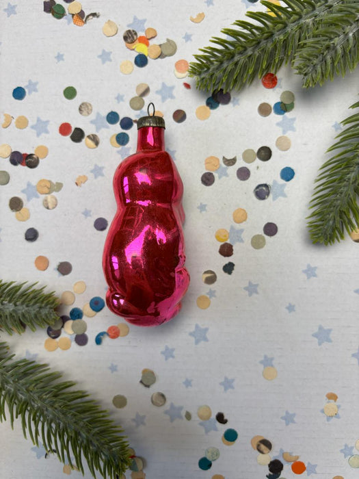 A shiny red ornament is placed on a white background, surrounded by colorful confetti and green pine branches.