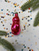 A shiny red ornament is placed on a white background, surrounded by colorful confetti and green pine branches.