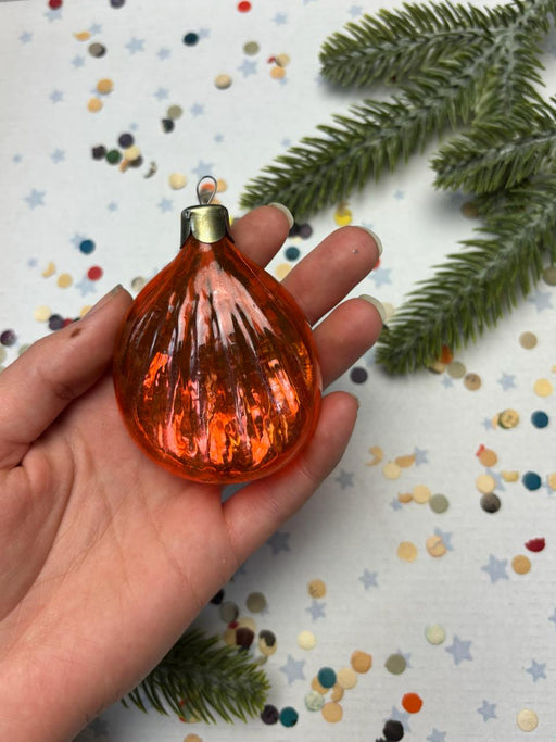 A hand holding a small orange glass ornament against a background of scattered confetti and a green pine branch.