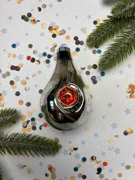 A shiny, metallic ornament with a red center is surrounded by colorful confetti on a white background, with a sprig of greenery to the side.