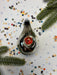 A shiny, metallic ornament with a red center is surrounded by colorful confetti on a white background, with a sprig of greenery to the side.