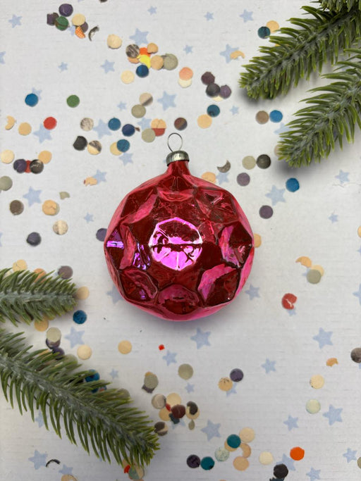 A red, diamond-shaped ornament with a glowing center is surrounded by scattered confetti and green pine branches on a white background.