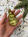 A hand holding a yellow glass ornament against a background of colorful confetti and a pine branch.