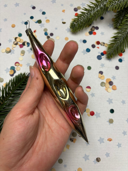 A hand is holding a metallic ornament with a pointed end, surrounded by colorful confetti and a pine tree branch.