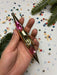 A hand is holding a metallic ornament with a pointed end, surrounded by colorful confetti and a pine tree branch.