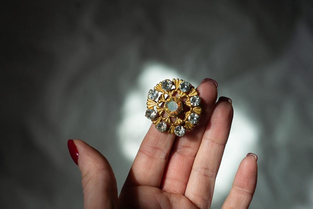 A close-up of a hand holding a decorative brooch with a central stone surrounded by smaller stones.