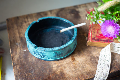 A blue ashtray with a lit cigarette sits on a wooden surface, accompanied by a book and a purple flower.