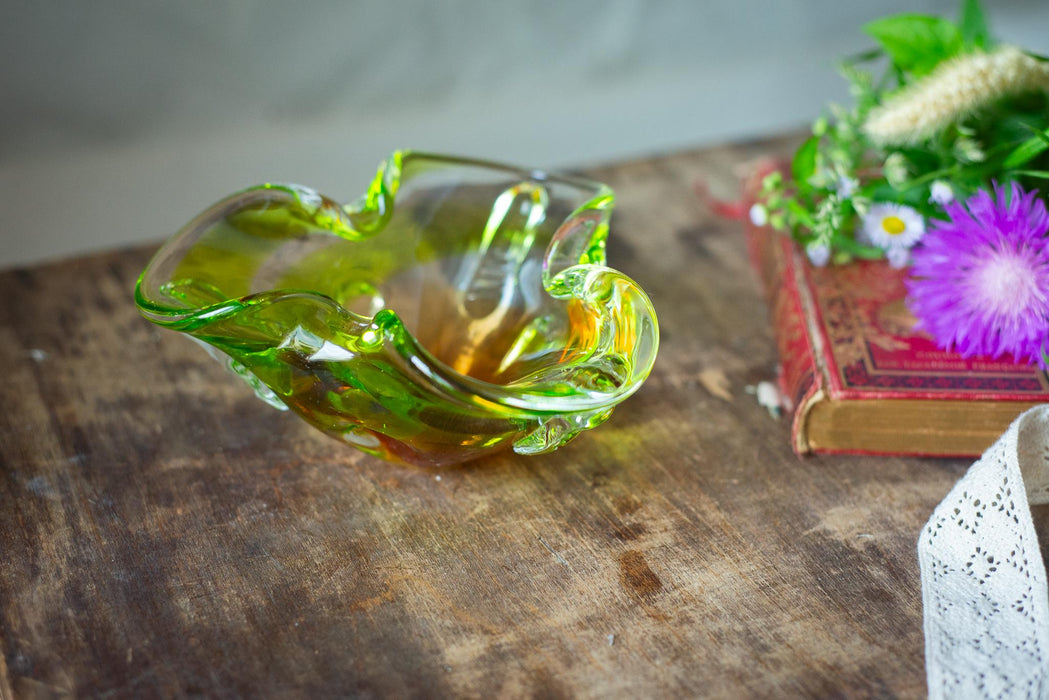 A green glass bowl with a curved shape sits on a wooden surface, accompanied by a bouquet of purple flowers and a red book.