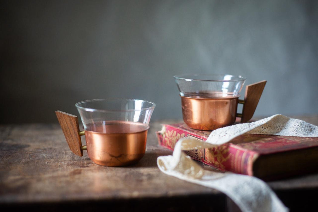 Two copper-colored mugs with wooden handles sit on a wooden surface, accompanied by a red and gold book and a white lace doily.