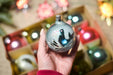 A hand holding a decorative ornament with a bird design, surrounded by various other ornaments in a box.