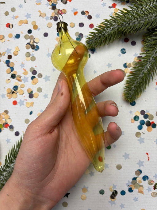 A hand holding a yellow ornament against a background of confetti and pine branches.