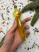 A hand holding a yellow ornament against a background of confetti and pine branches.