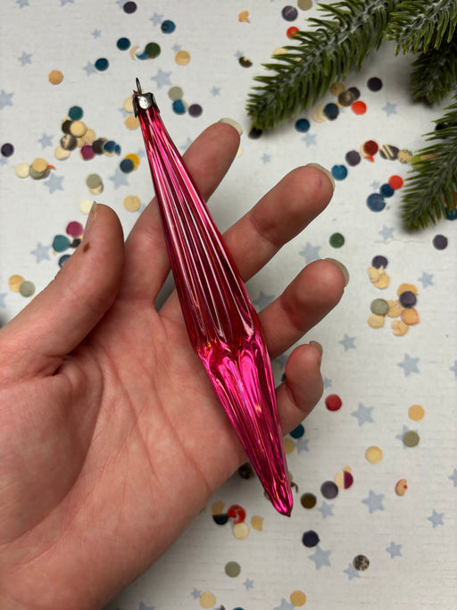 A hand holding a pink glass ornament against a background of colorful confetti and a pine tree branch.